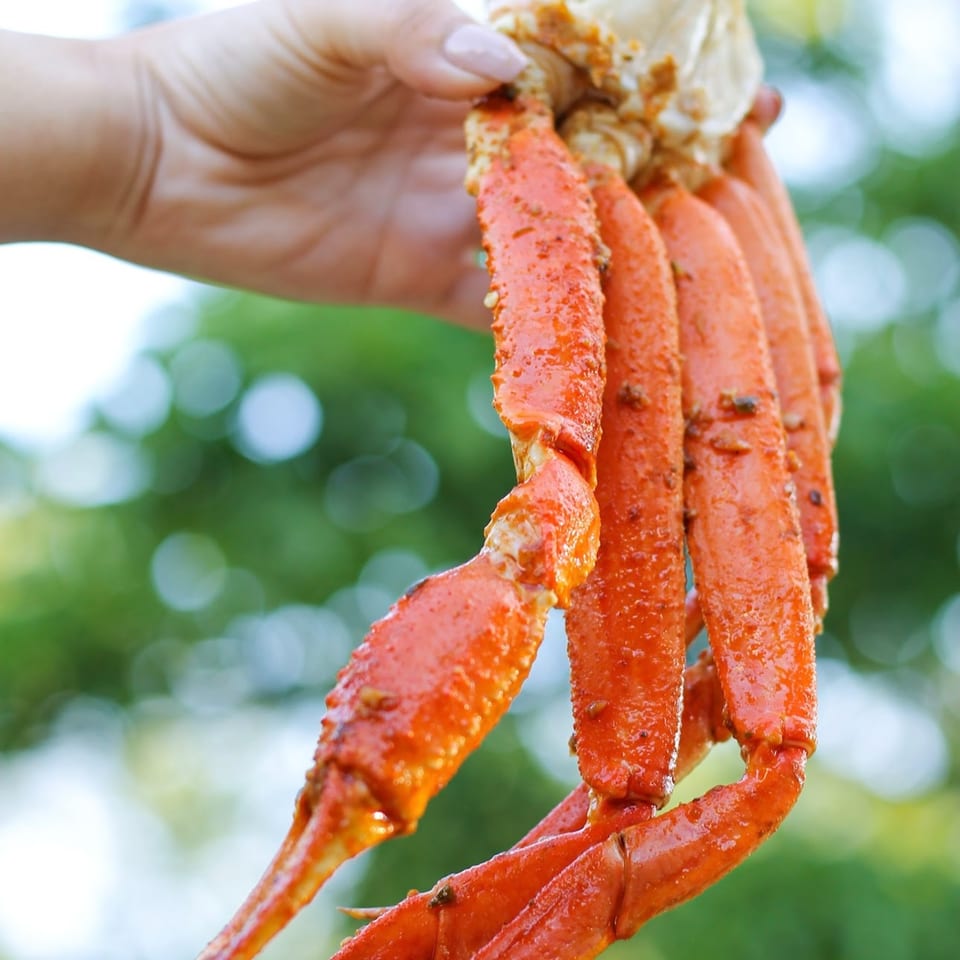 A hand holds up several bright orange, seasoned crab legs against a blurred green background.