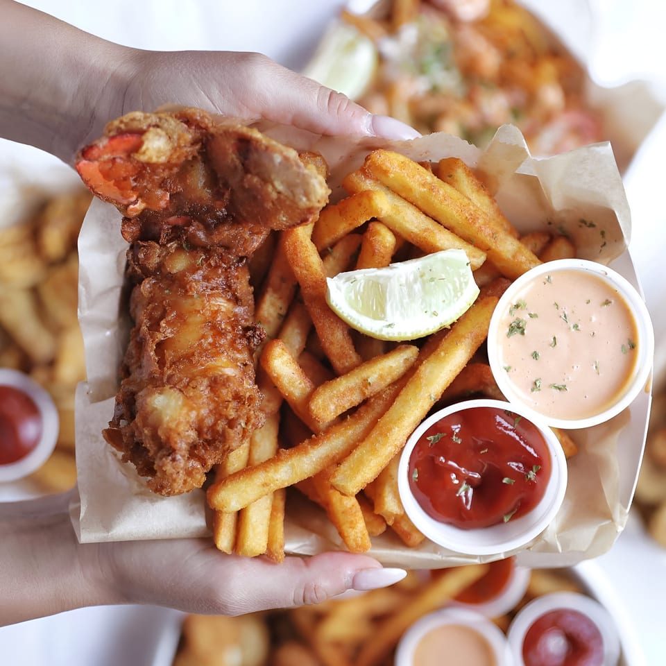 Hands holding a Shaking Crab seafood basket with fried lobster, fries, lemon, ketchup, and sauce.
