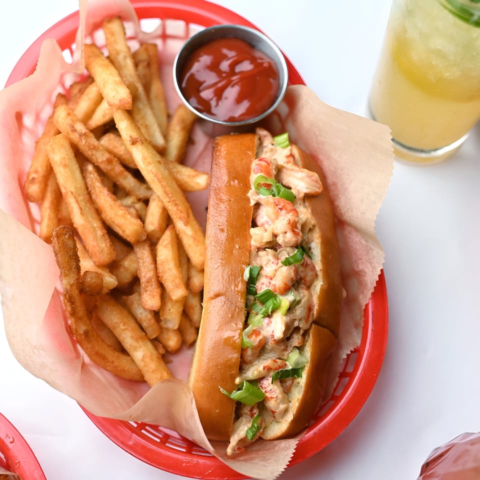 A Shaking Crab lobster roll with fries and ketchup in a red basket, with a drink beside it.